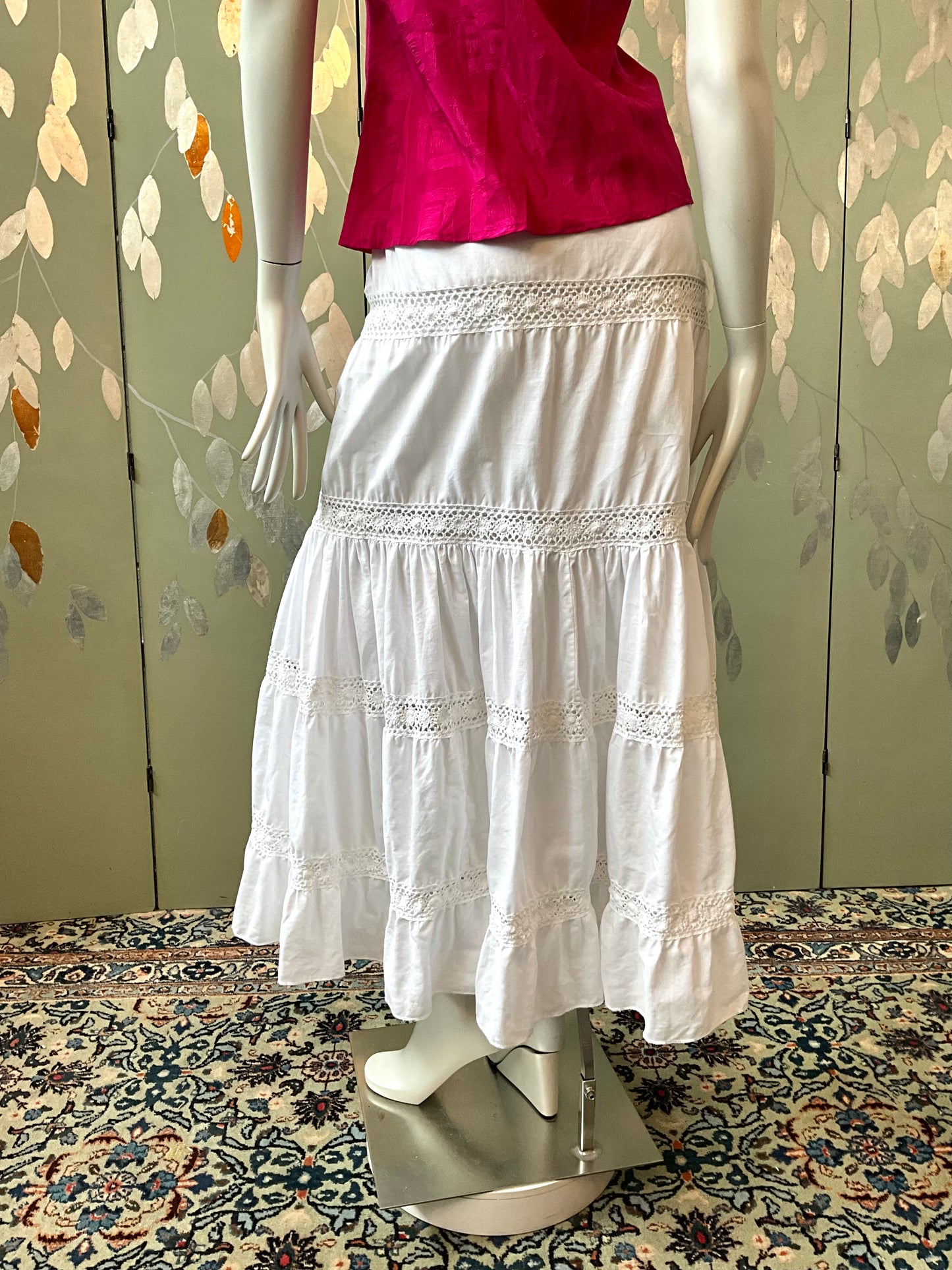 Mannequin wearing a pink top and white skirt on a patterned rug with a floral wall background.