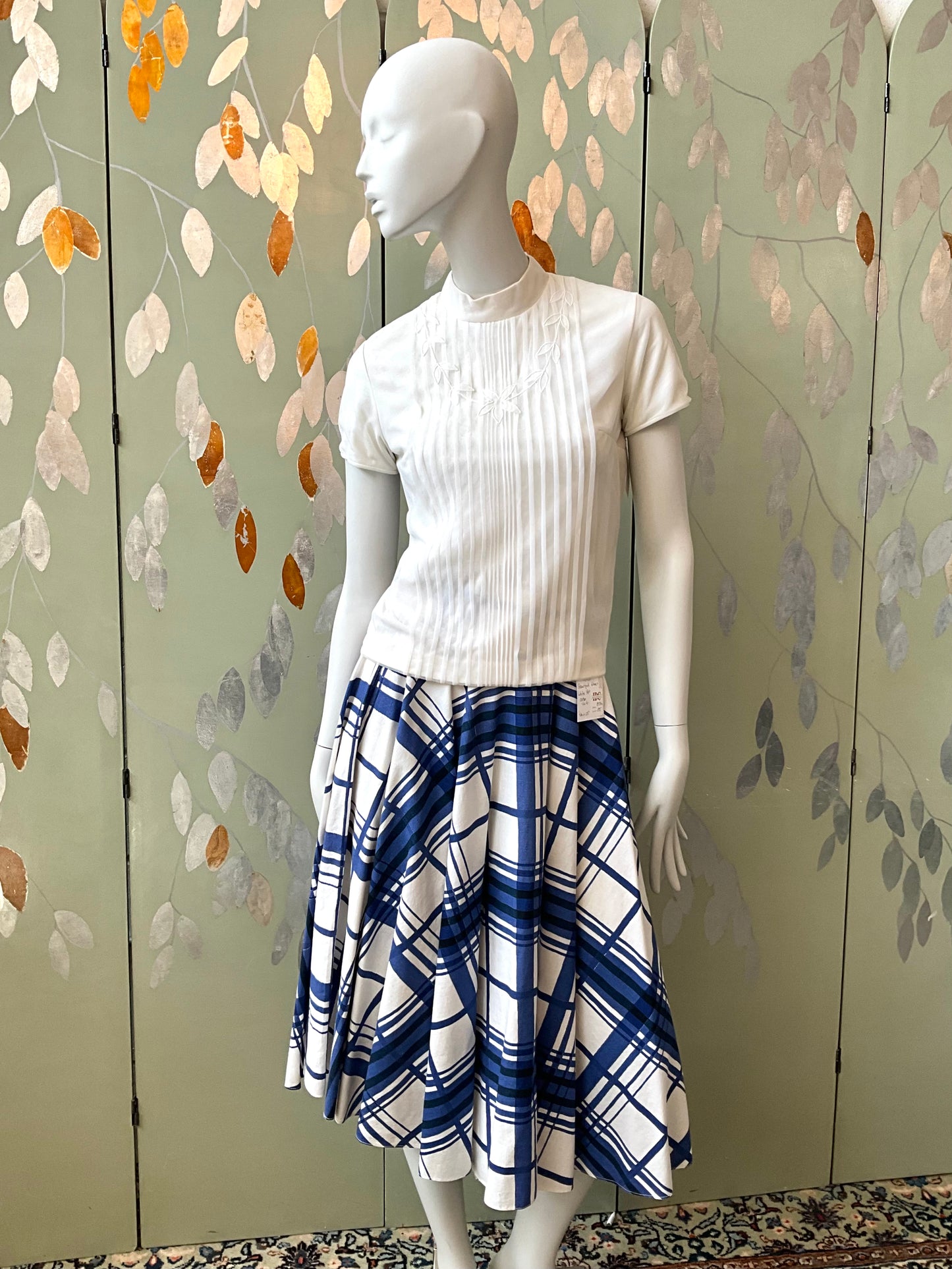 Mannequin wearing a white top and blue plaid skirt against a decorative wall.