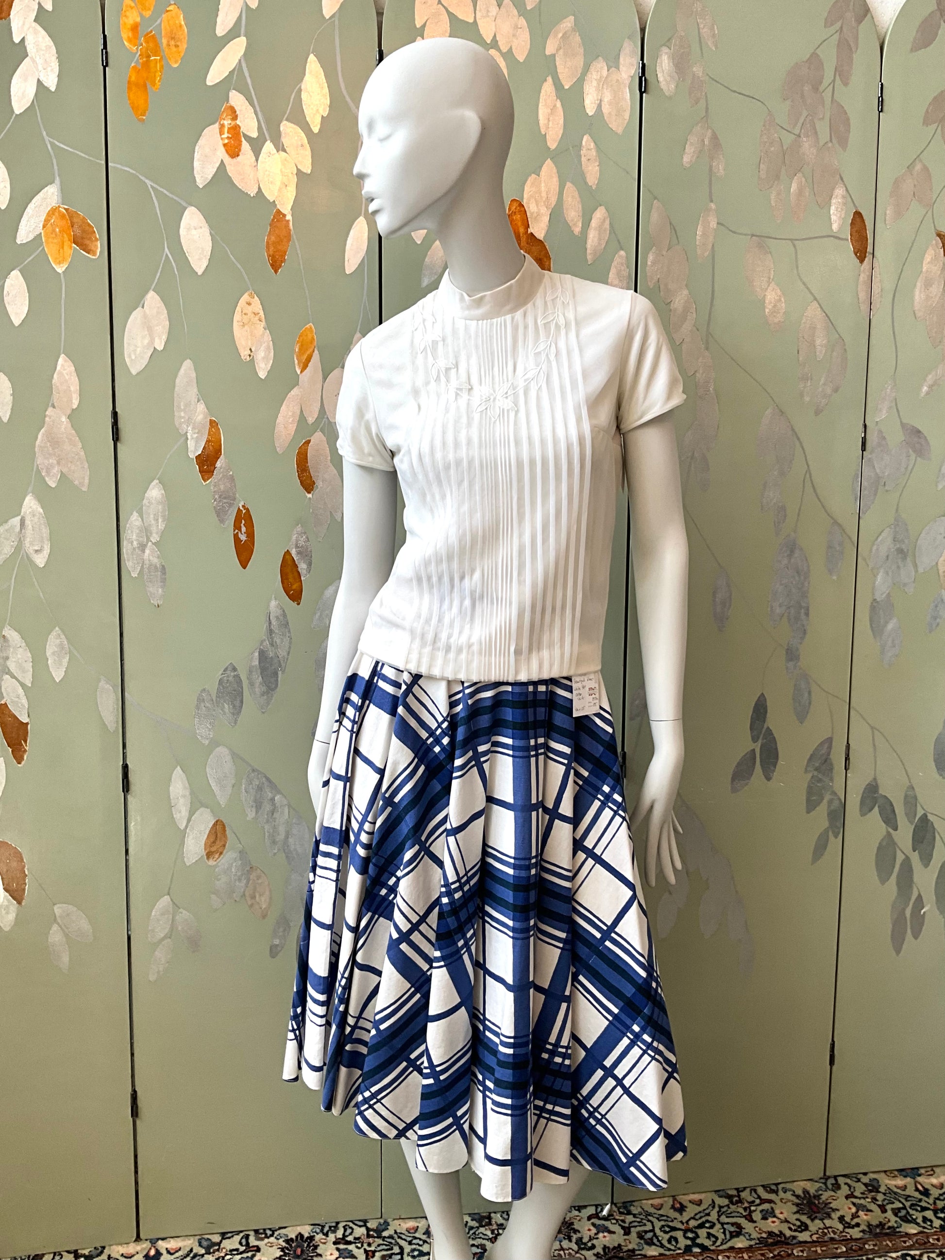 Mannequin wearing a white top and blue plaid skirt against a decorative wall.