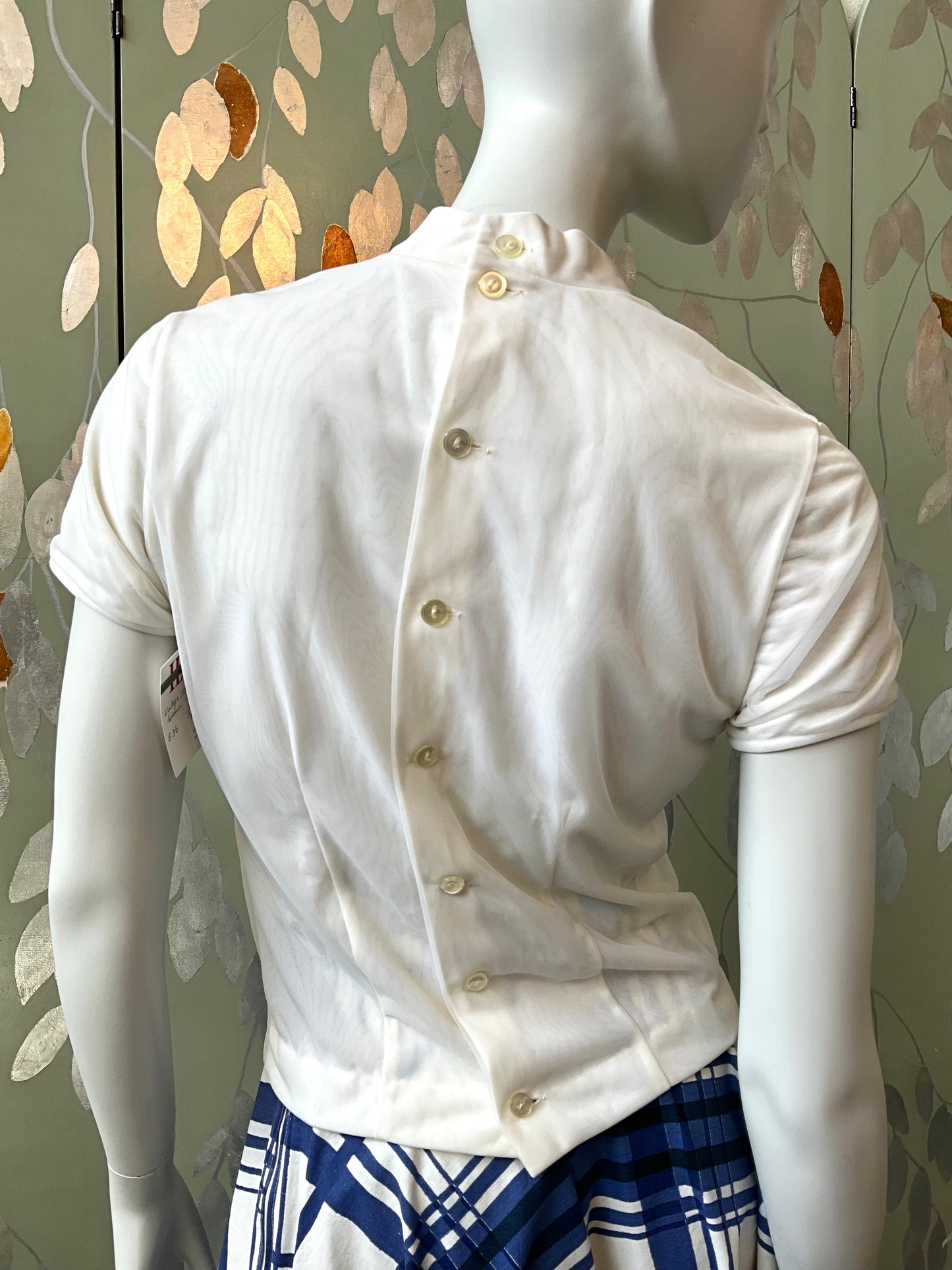 Mannequin wearing a white short-sleeve shirt with buttons against a decorative wall.