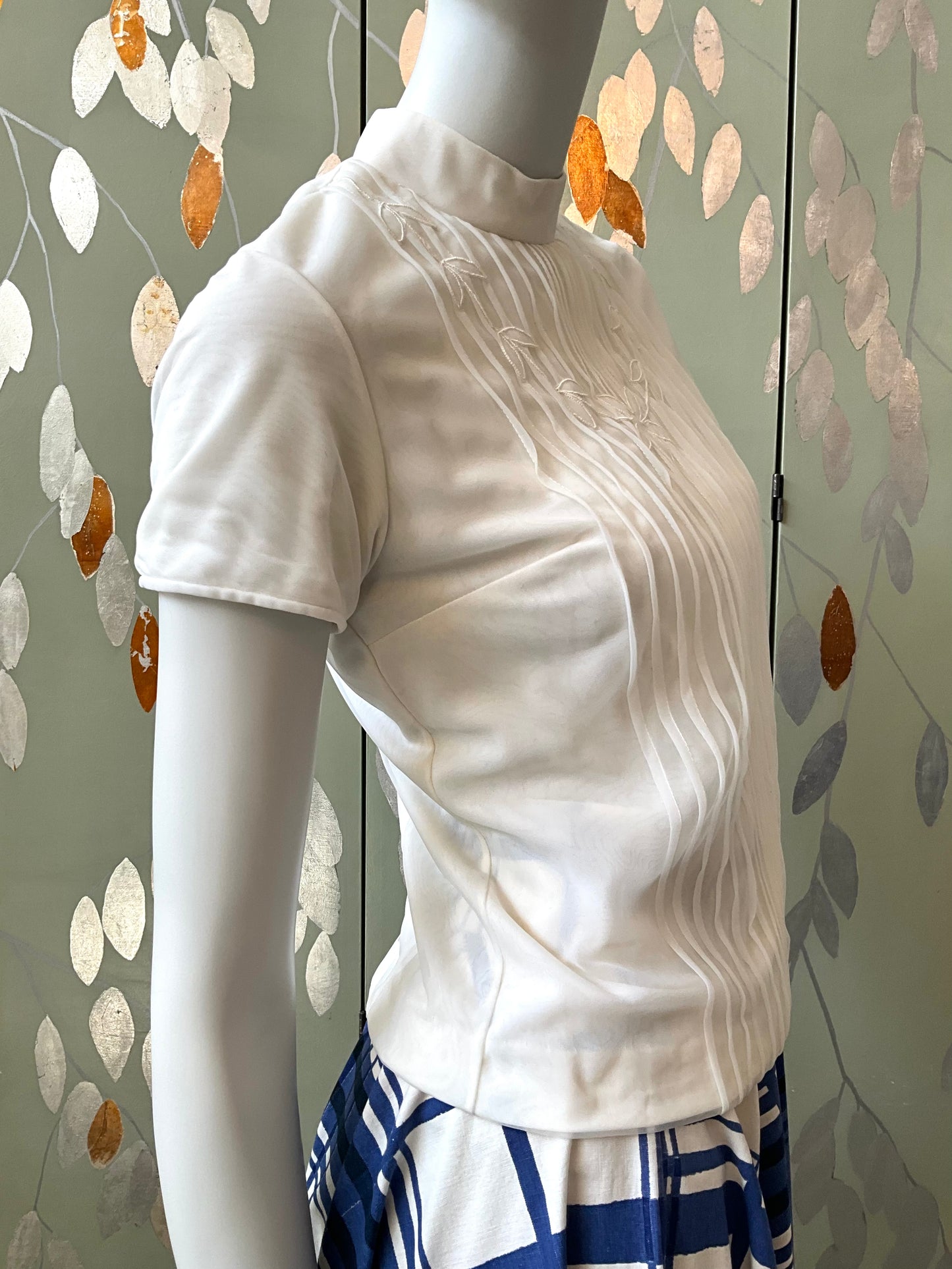 White short-sleeve top on a mannequin against a decorative wall.